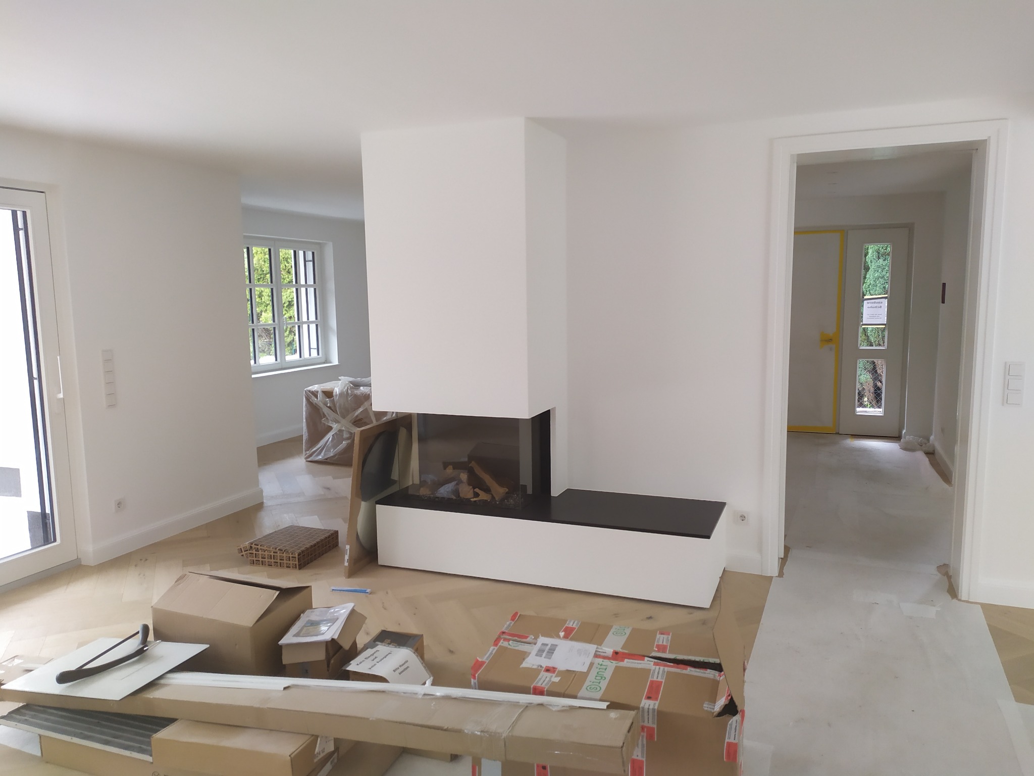 Wnętrze w trakcie prac wykończeniowych, z nowoczesnym kominkiem obudowanym białą konstrukcją z czarnym blatem, kartony i narzędzia na podłodze z jasnego drewna, widok na kolejne pomieszczenia przez...
