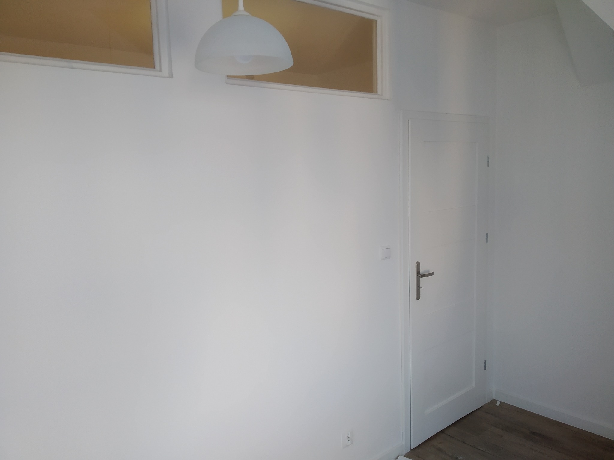 Świeżo pomalowane na biało wnętrze pokoju z białymi ścianami, białymi drzwiami z srebrną klamką i drewnianą podłogą, oświetlone lampą wiszącą i światłem wpadającym przez okno.