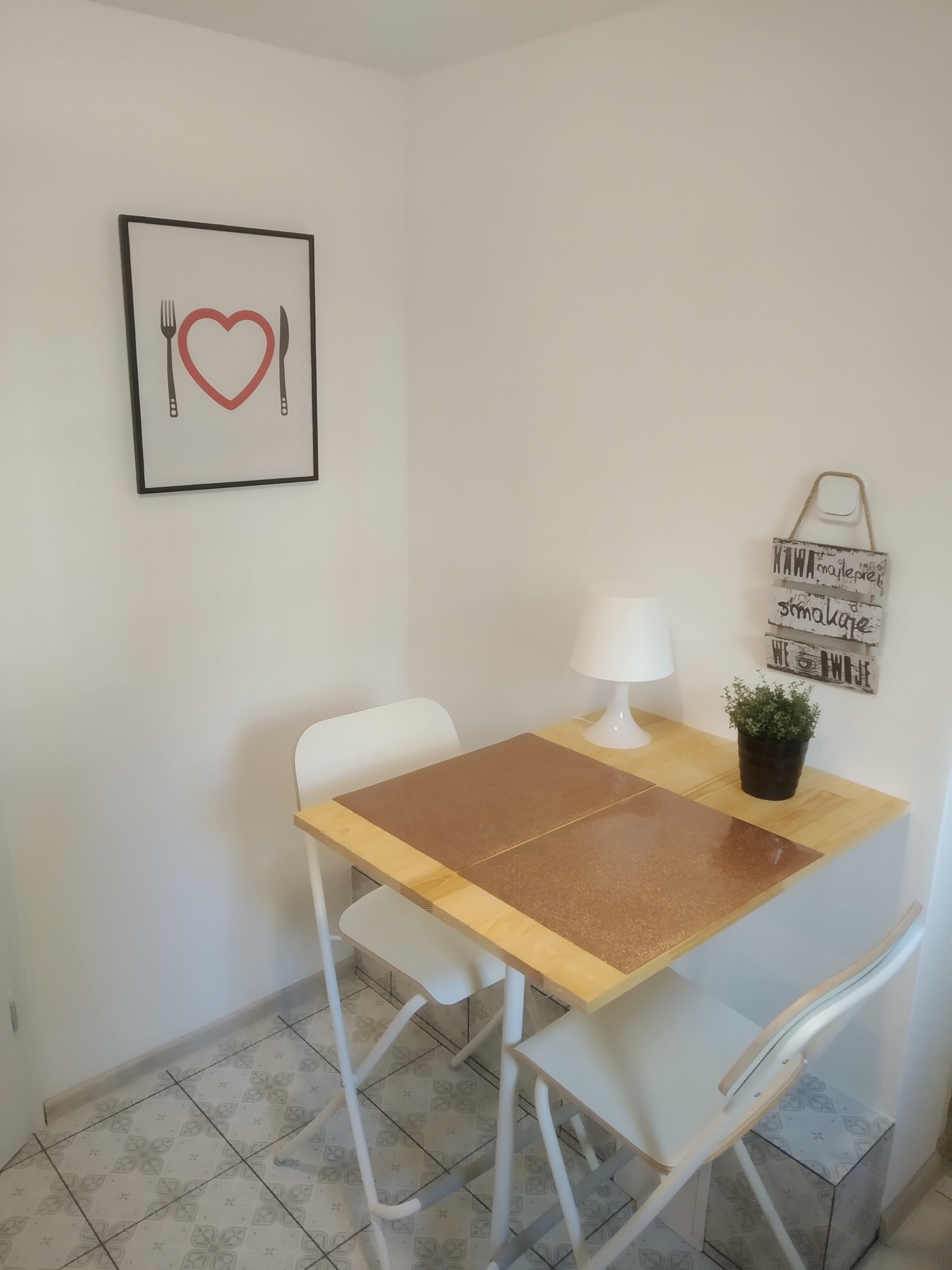 Narożnik jadalny z drewnianym stołem, białymi krzesłami składanymi, lampką i rośliną w doniczce, dekorowany obrazkiem z sercem i napisem 'Kawa smakuje najlepiej we dwoje'.