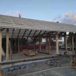 Drewniana konstrukcja z zadaszeniem w trakcie budowy. Widoczne narzędzia i materiały budowlane. W tle dom i zachmurzone niebo.