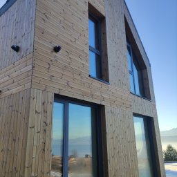 Jakub Bednarczyk budownictwo drewniane - Nowoczesny dom z elewacją z jasnego drewna, okna z ciemnymi ramami, widok na góry w tle. Ujęcie z dołu, podkreślające geometrię budynku.