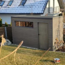 Nowoczesna, szara, drewniana konstrukcja z oknem i drzwiami w ogrodzie, na tle domu z oknami dachowymi i śniegiem na dachu. W trawie widoczny zwinięty kabel.