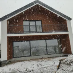 Nowoczesny dom dwukondygnacyjny z drewnianą elewacją i balkonem z metalową balustradą, sfotografowany podczas śnieżycy, z widocznymi śladami budowy na zewnątrz.