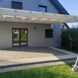 Nowoczesna pergola z jasnego drewna nad tarasem wyłożonym szarą kostką brukową, przylegająca do szarego domu z brązowymi oknami i drzwiami.