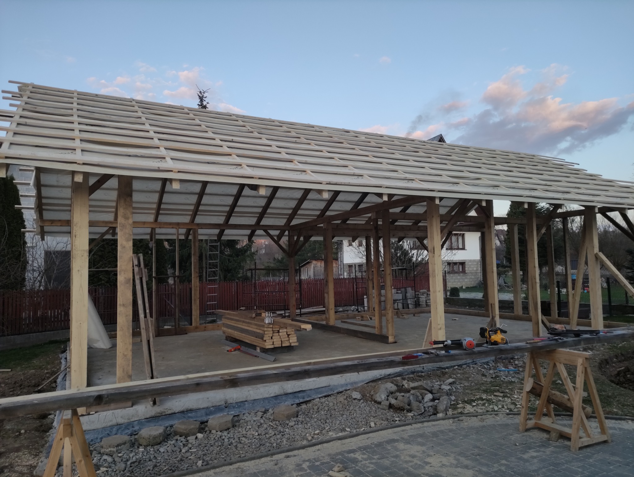 Drewniana konstrukcja z zadaszeniem w trakcie budowy. Widoczne narzędzia i materiały budowlane. W tle dom i zachmurzone niebo.