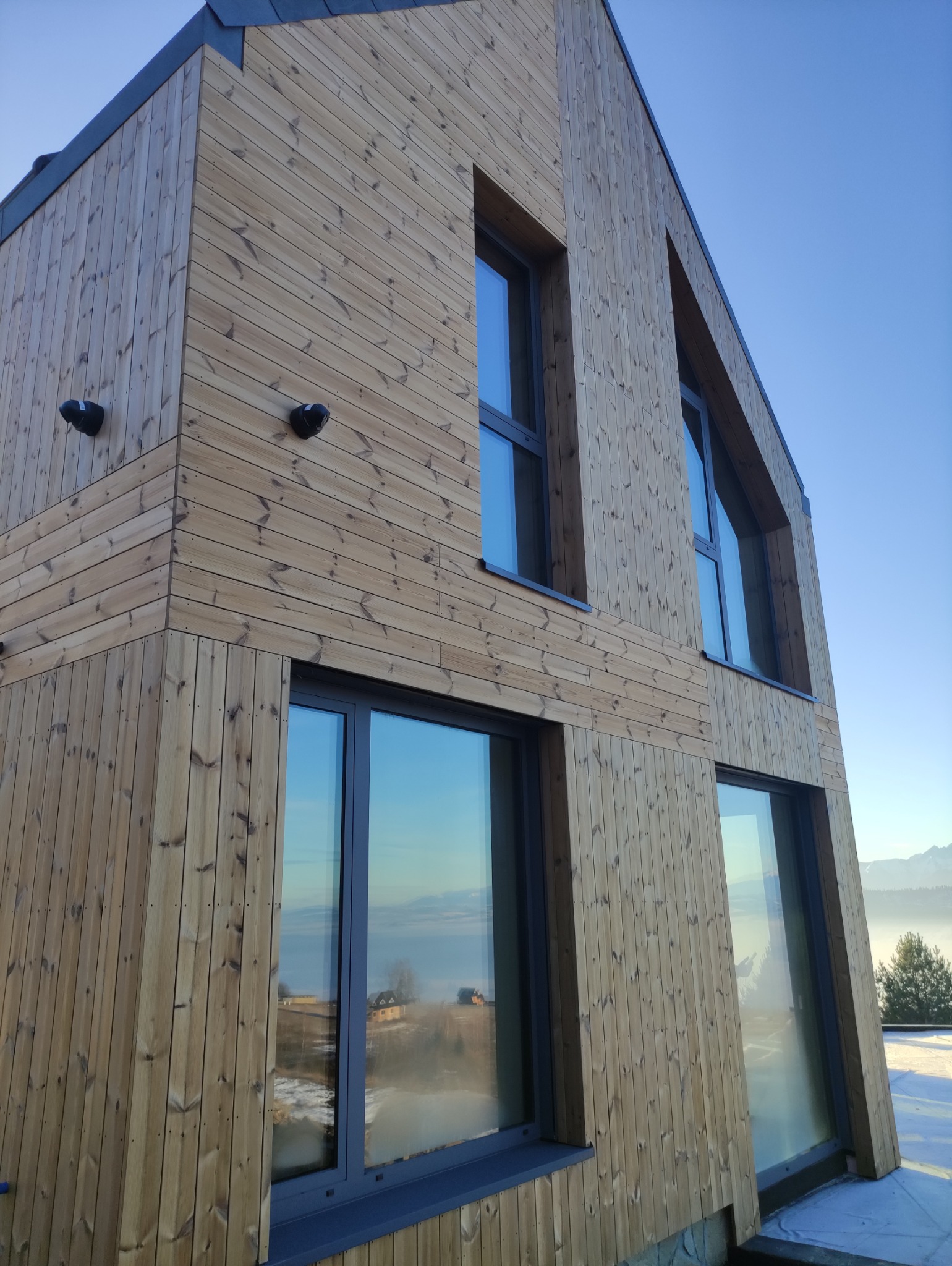 Nowoczesny dom z elewacją z jasnego drewna, okna z ciemnymi ramami, widok na góry w tle. Ujęcie z dołu, podkreślające geometrię budynku.