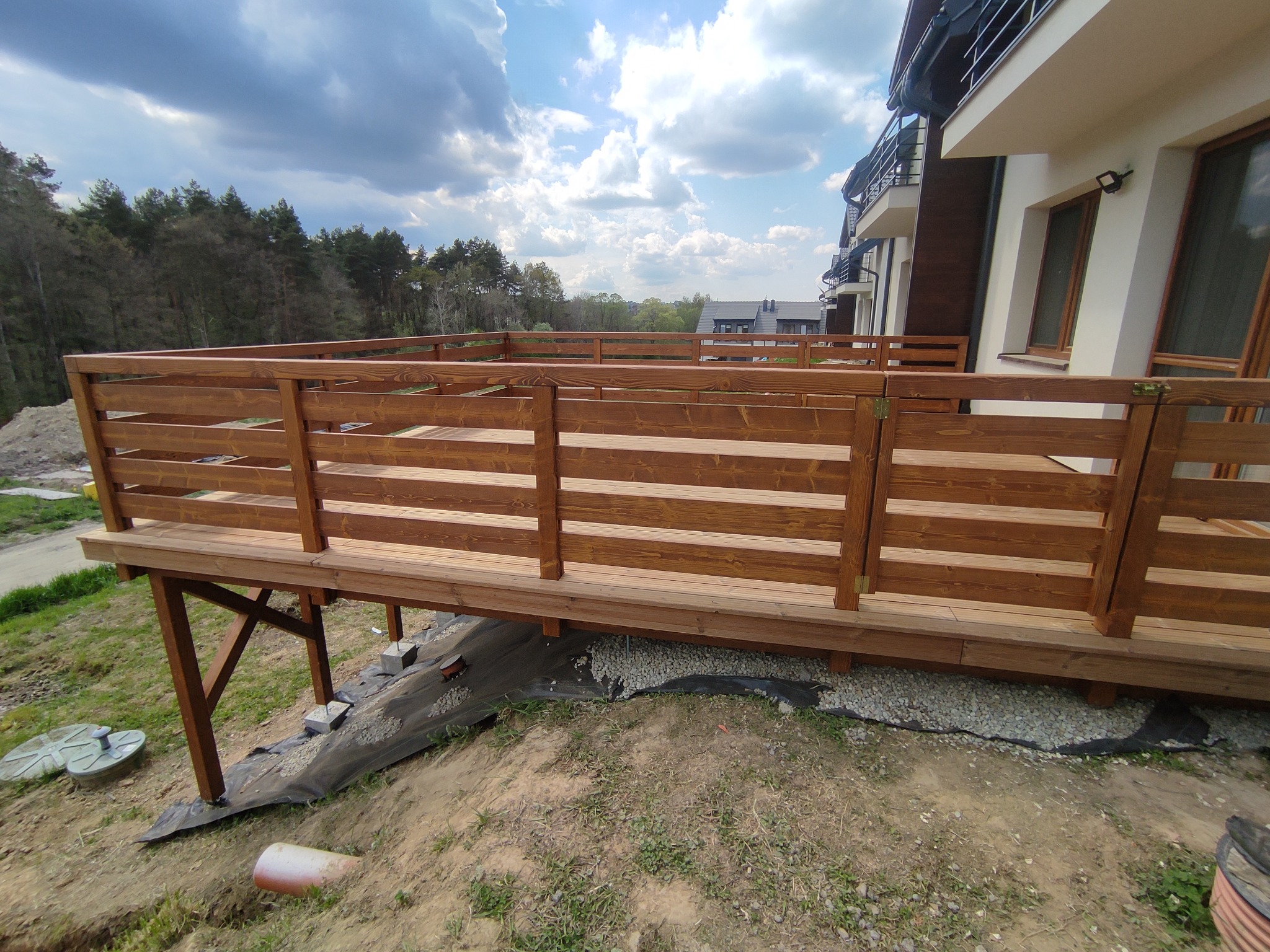 Nowo wybudowany taras drewniany z balustradą, widok z boku, z widocznymi elementami konstrukcyjnymi i przygotowanym podłożem z geowłókniny i kamienia.