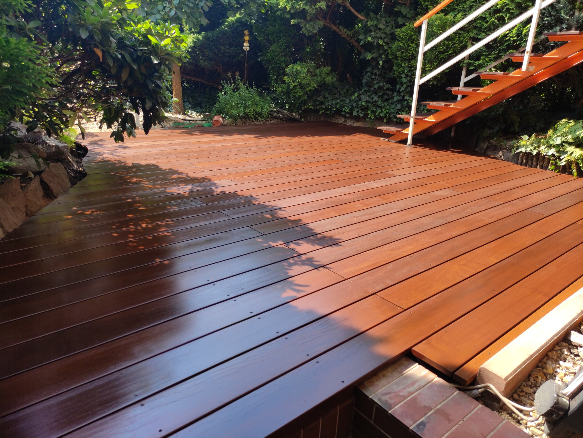 Świeżo położony, brązowy taras z drewna egzotycznego, widoczne schody z białymi poręczami, cień drzew na deskach, bujna roślinność w tle.