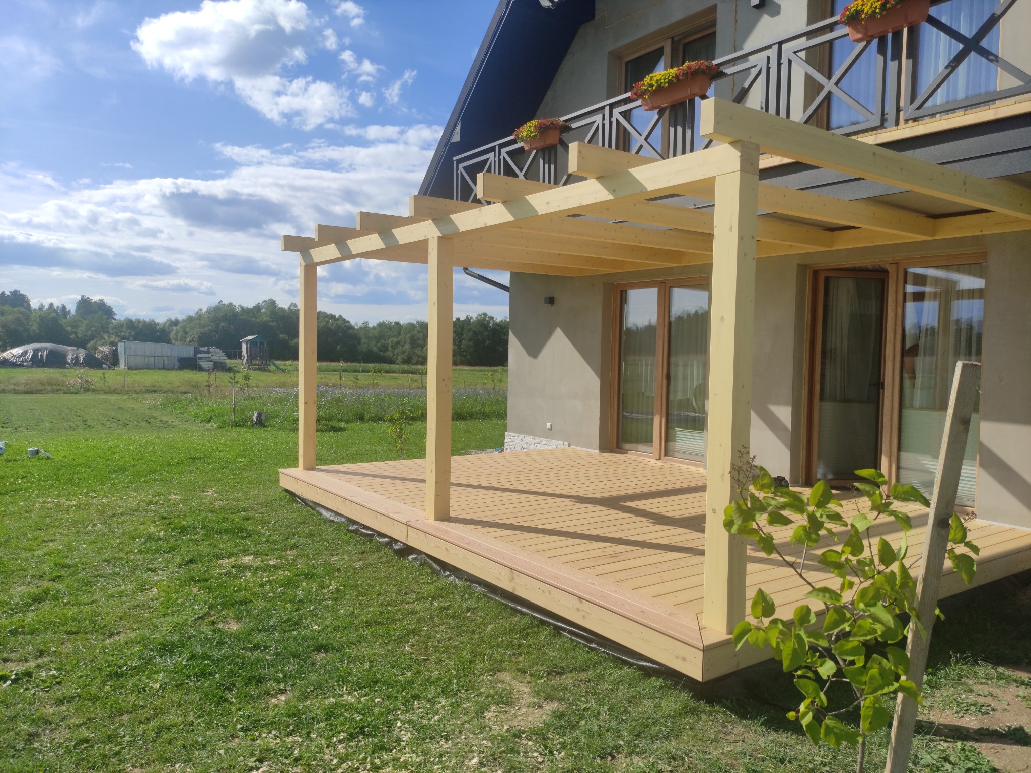 Jasny, drewniany taras z pergolą przylegający do domu z balkonem, widok na zielony trawnik i odległe zabudowania gospodarcze pod błękitnym, lekko zachmurzonym niebem.
