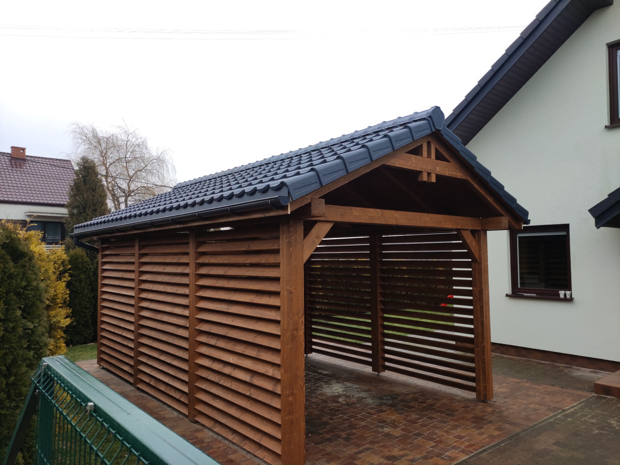 Drewniana wiata garażowa z lamelowymi ścianami i ciemną dachówką, przylegająca do domu z jasną elewacją, na wybrukowanym podjeździe.