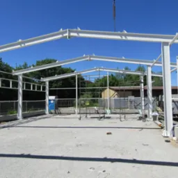 Konstrukcja stalowa hali w trakcie montażu, widoczny dźwig i rusztowania na placu budowy, błękitne niebo w tle.