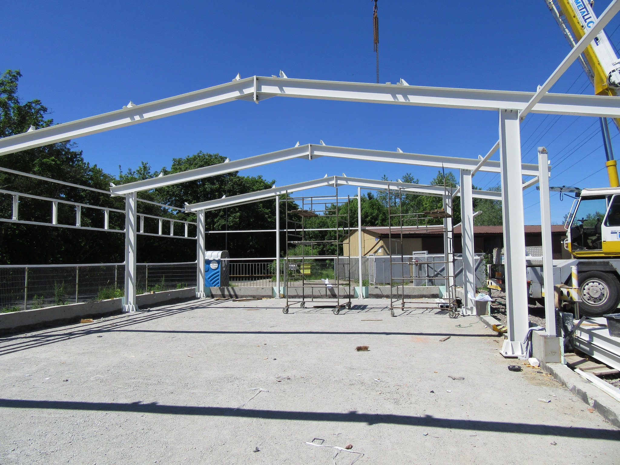 Konstrukcja stalowa hali w trakcie montażu, widoczny dźwig i rusztowania na placu budowy, błękitne niebo w tle.