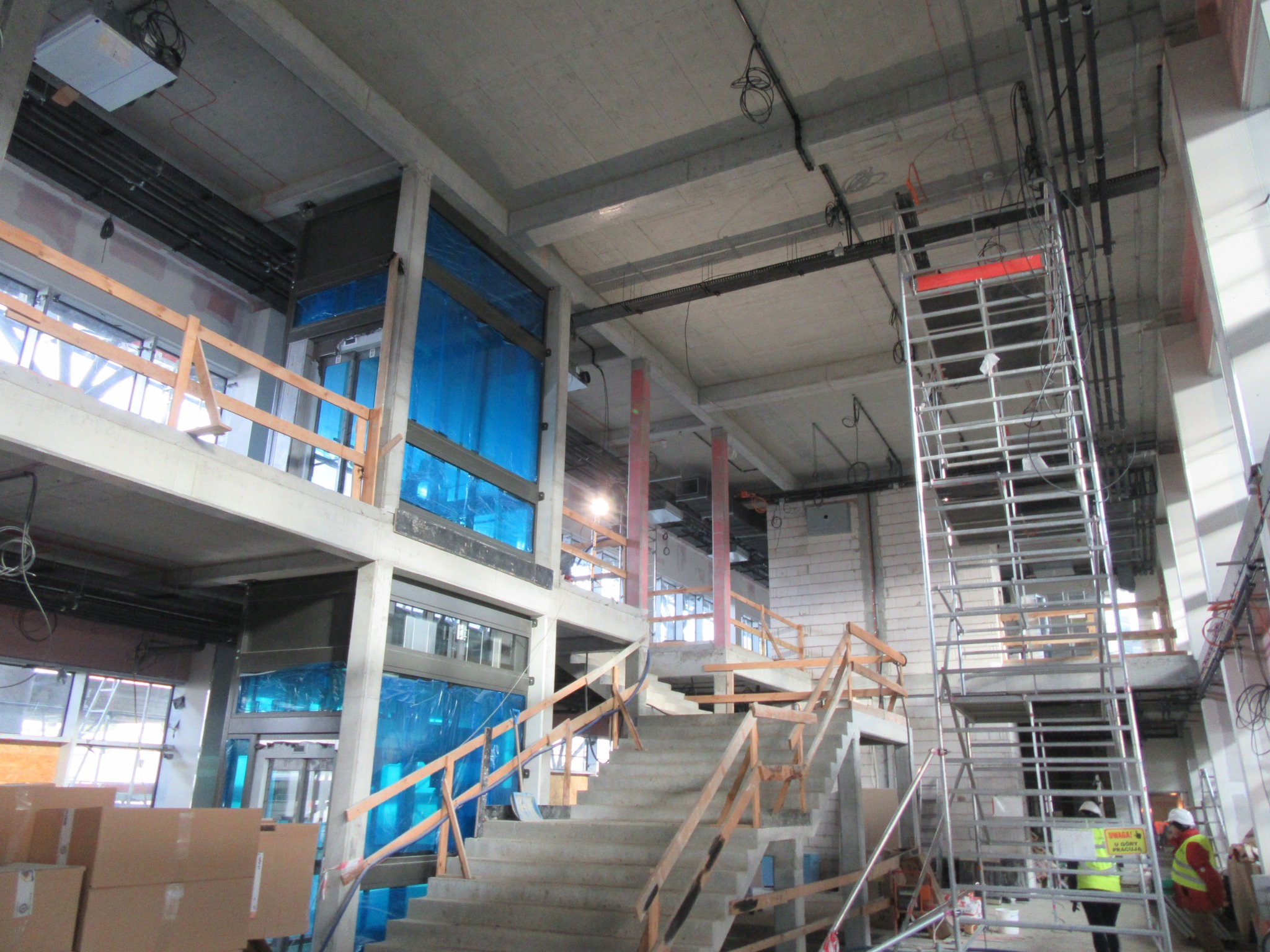 Wnętrze surowego budynku w trakcie budowy: betonowe schody z drewnianą barierką, aluminiowe rusztowanie, okna zabezpieczone folią, widoczne instalacje elektryczne pod sufitem, pracownicy...