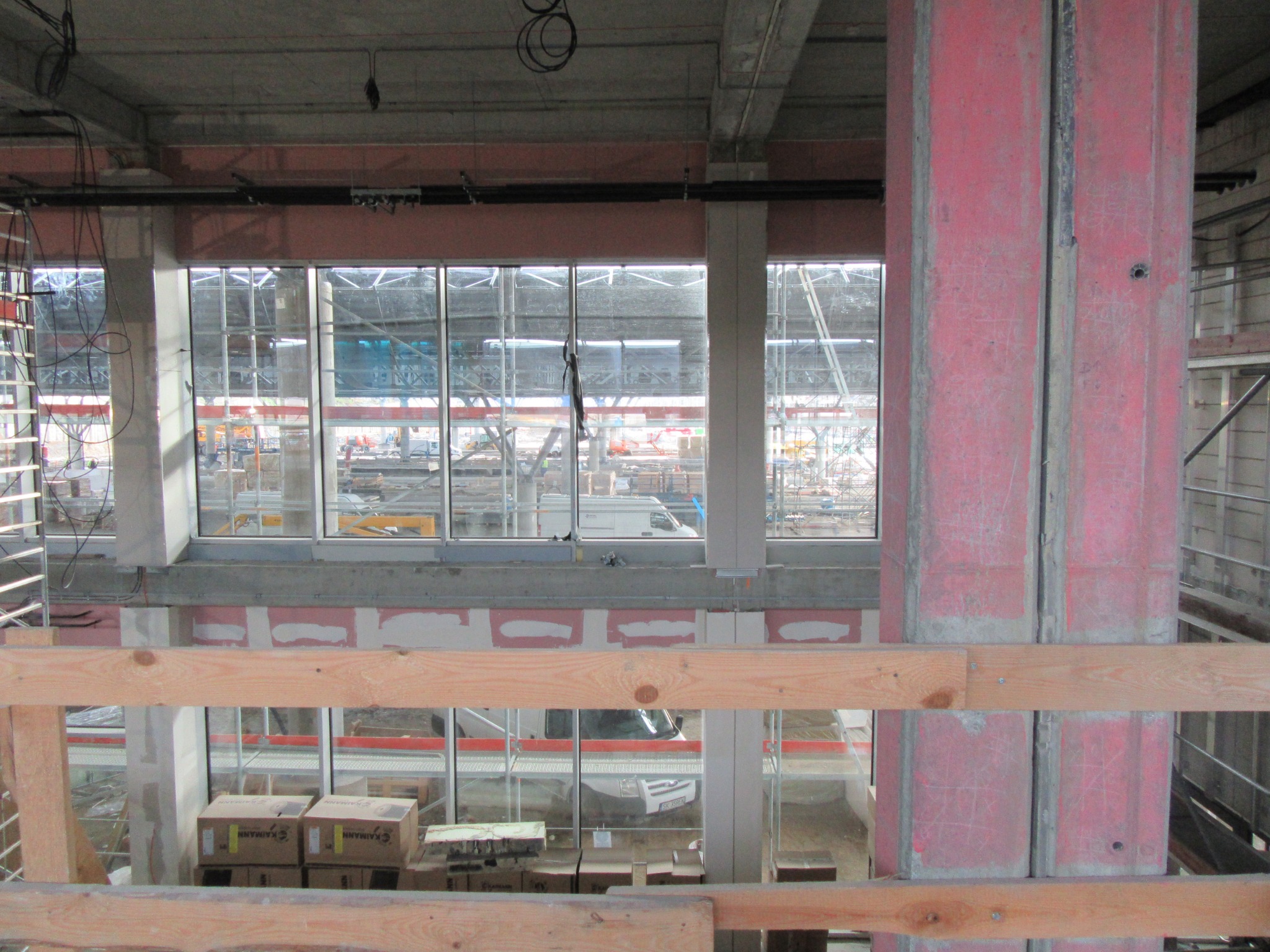 Wnętrze budynku w stanie surowym: widoczne betonowe ściany, metalowe słupy pomalowane na czerwono, okna z widokiem na plac budowy, drewniane elementy konstrukcyjne i kartony na podłodze.