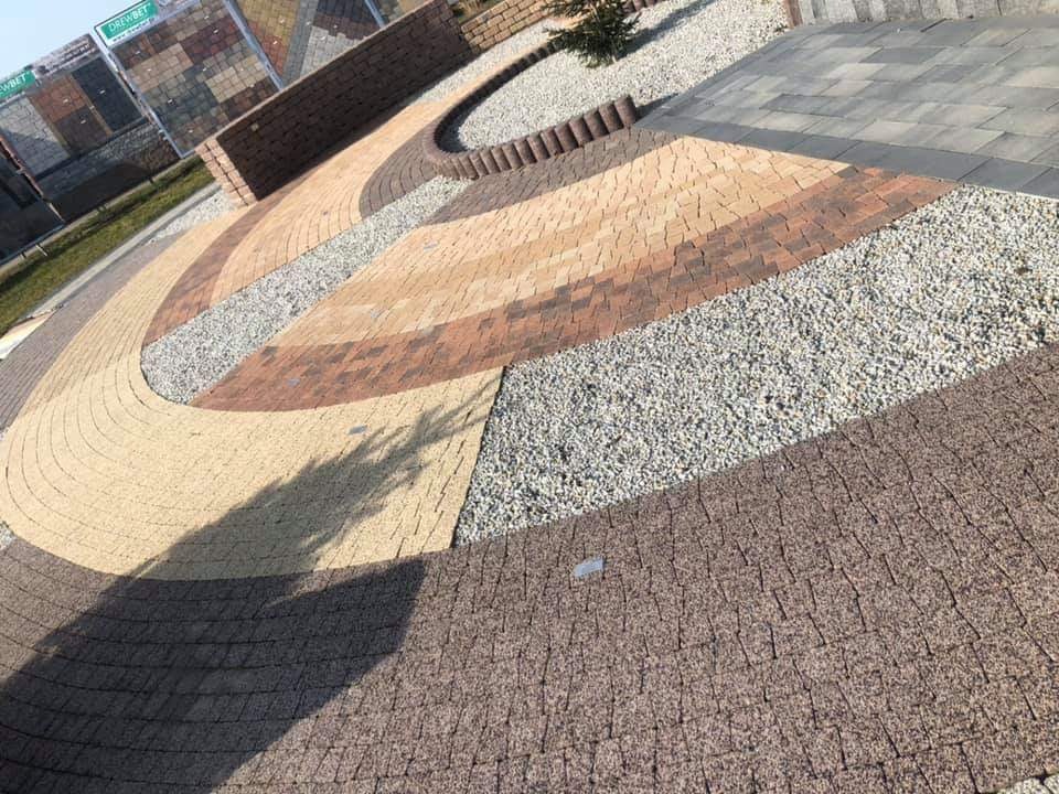 Wzorcowe ułożenie kostki brukowej w odcieniach brązu i beżu, oddzielonej pasami żwiru, prezentowane na ekspozycji zewnętrznej z widocznym logo producenta kostki w tle.