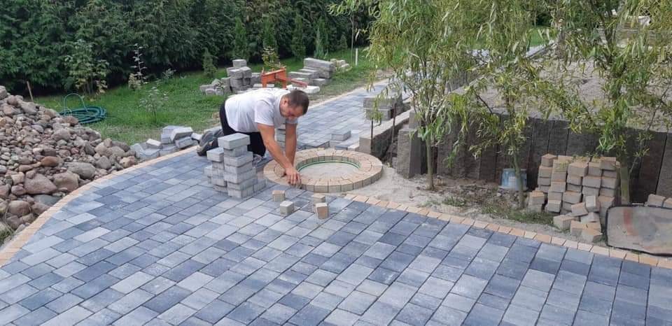 Pracownik w trakcie tworzenia okrągłego elementu z jasnej kostki na tle ułożonej z szarej kostki ścieżki ogrodowej, widoczne stosy kostki i otoczenie zieleni.