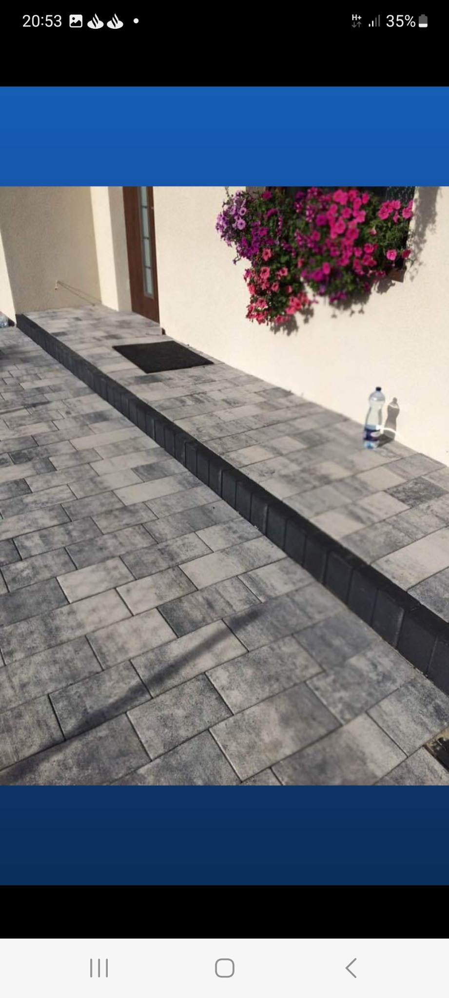 Ułożona szara kostka brukowa prowadząca do drzwi wejściowych, obok ściany z kwiatami w doniczce, widoczna butelka wody mineralnej.