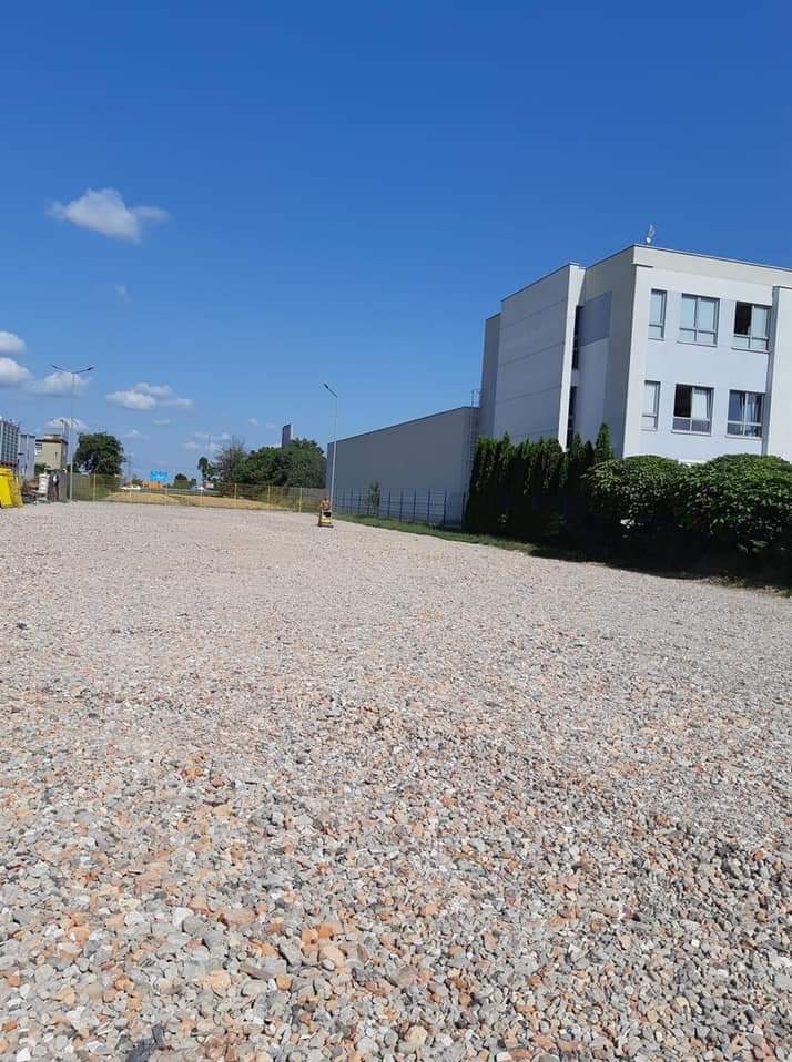 Szeroki, żwirowy plac budowy z widocznym budynkiem w tle, oświetlony jasnym, dziennym światłem, z żółtą maszyną budowlaną w oddali.