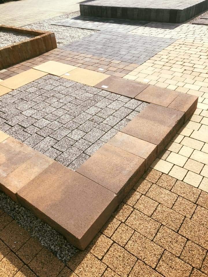 Ekspozycja różnych wzorów i kolorów kostki brukowej na placu wystawowym, ukazująca kwadrat z szarej kostki granitowej otoczony brązową obwódką, sąsiadujący z żółtą kostką brukową i szarym brukiem...