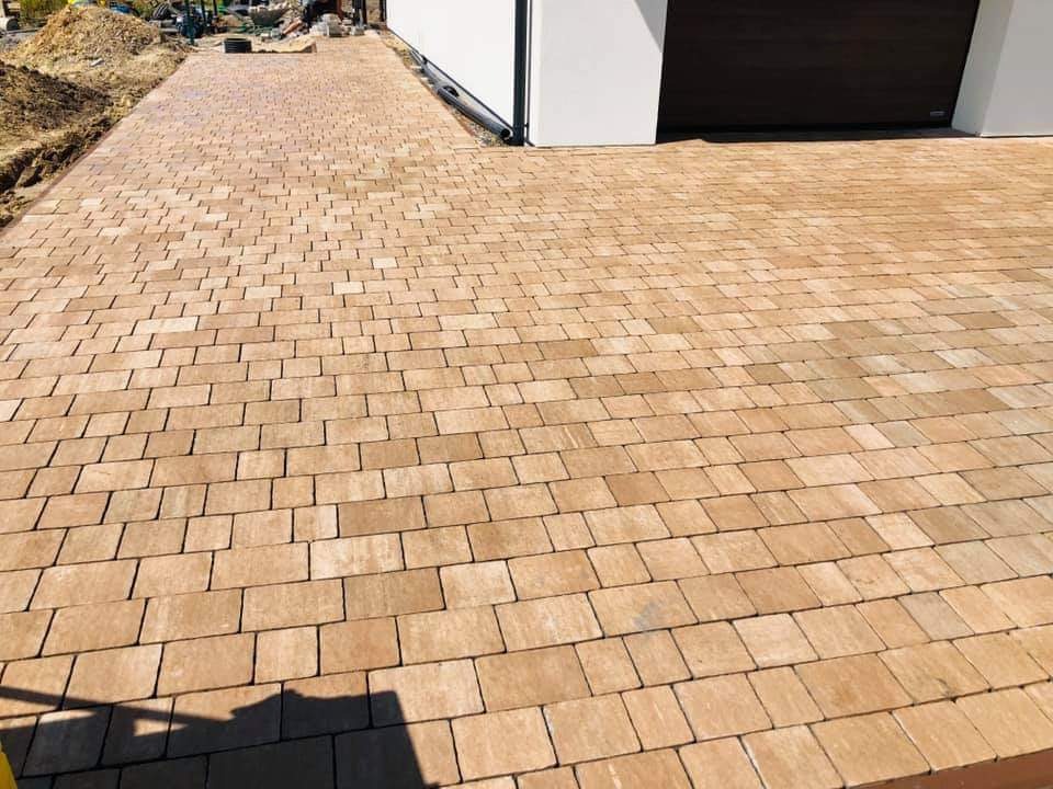 Ułożona, beżowa kostka brukowa prowadząca do garażu z brązową bramą, widoczne ślady piasku i ziemi po bokach.