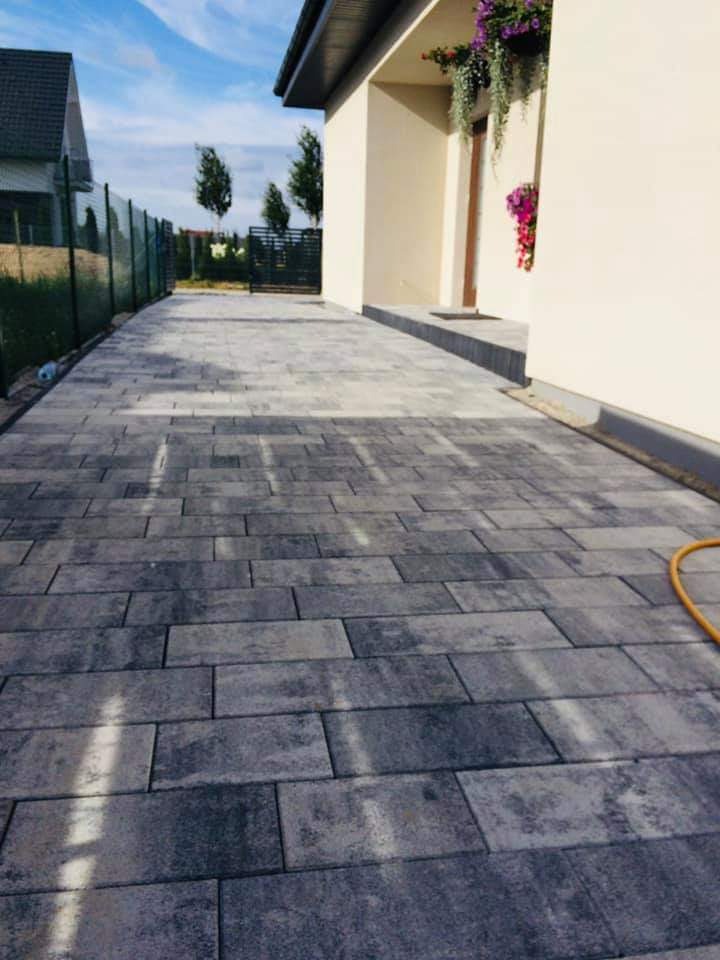 Ułożona szara kostka brukowa przed domem jednorodzinnym z kwiatami w donicach przy wejściu, widoczny fragment węża ogrodowego.