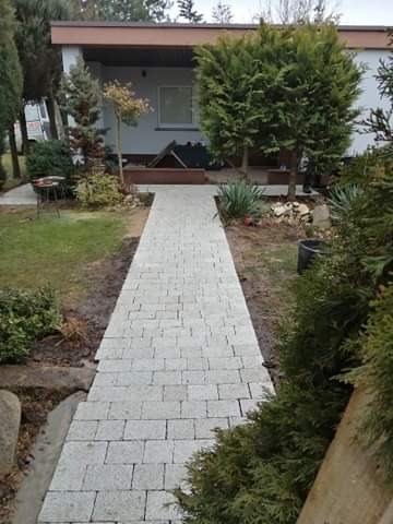 Ścieżka z jasnej kostki prowadząca do domu jednorodzinnego z brązowym dachem, otoczona zielenią i niskimi krzewami.