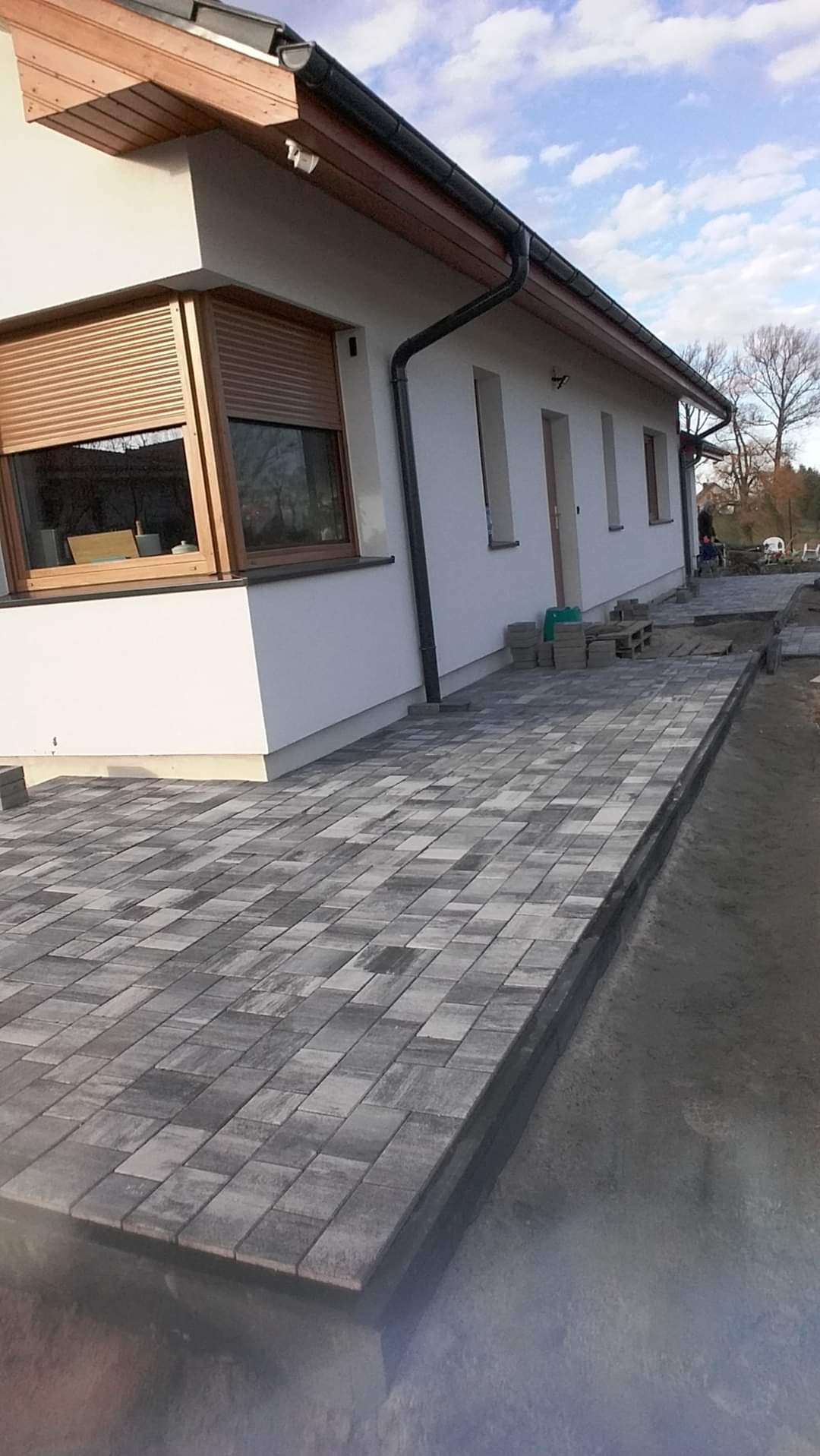 Ułożona, szara kostka brukowa tworząca chodnik przy białym budynku w Poznaniu, widoczne okno z brązowymi roletami i przygotowane do ułożenia kostki.