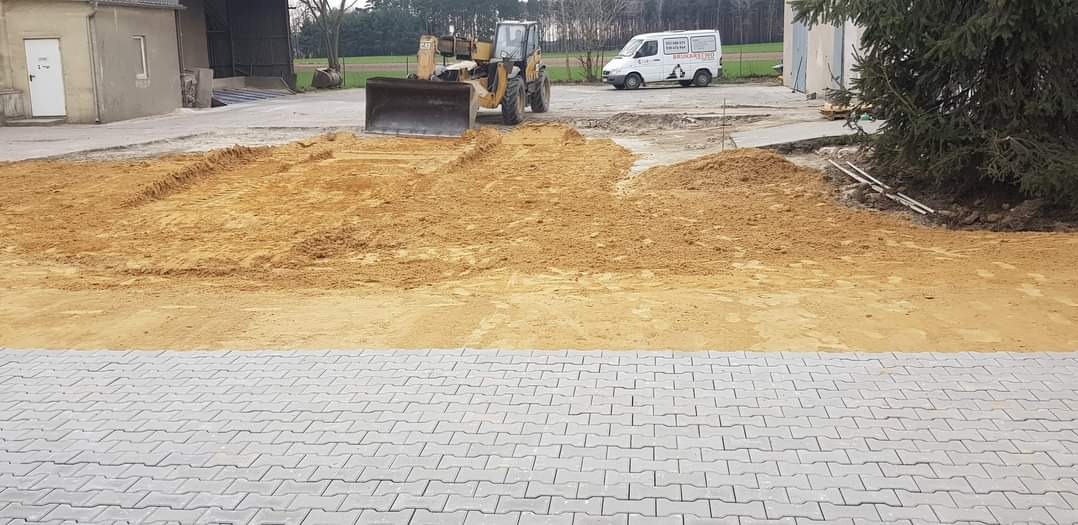 Żółta koparka z łyżką na placu budowy, przygotowującym teren pod układanie szarej kostki brukowej; w tle budynek gospodarczy, biały bus z reklamą firmy i drzewo iglaste.