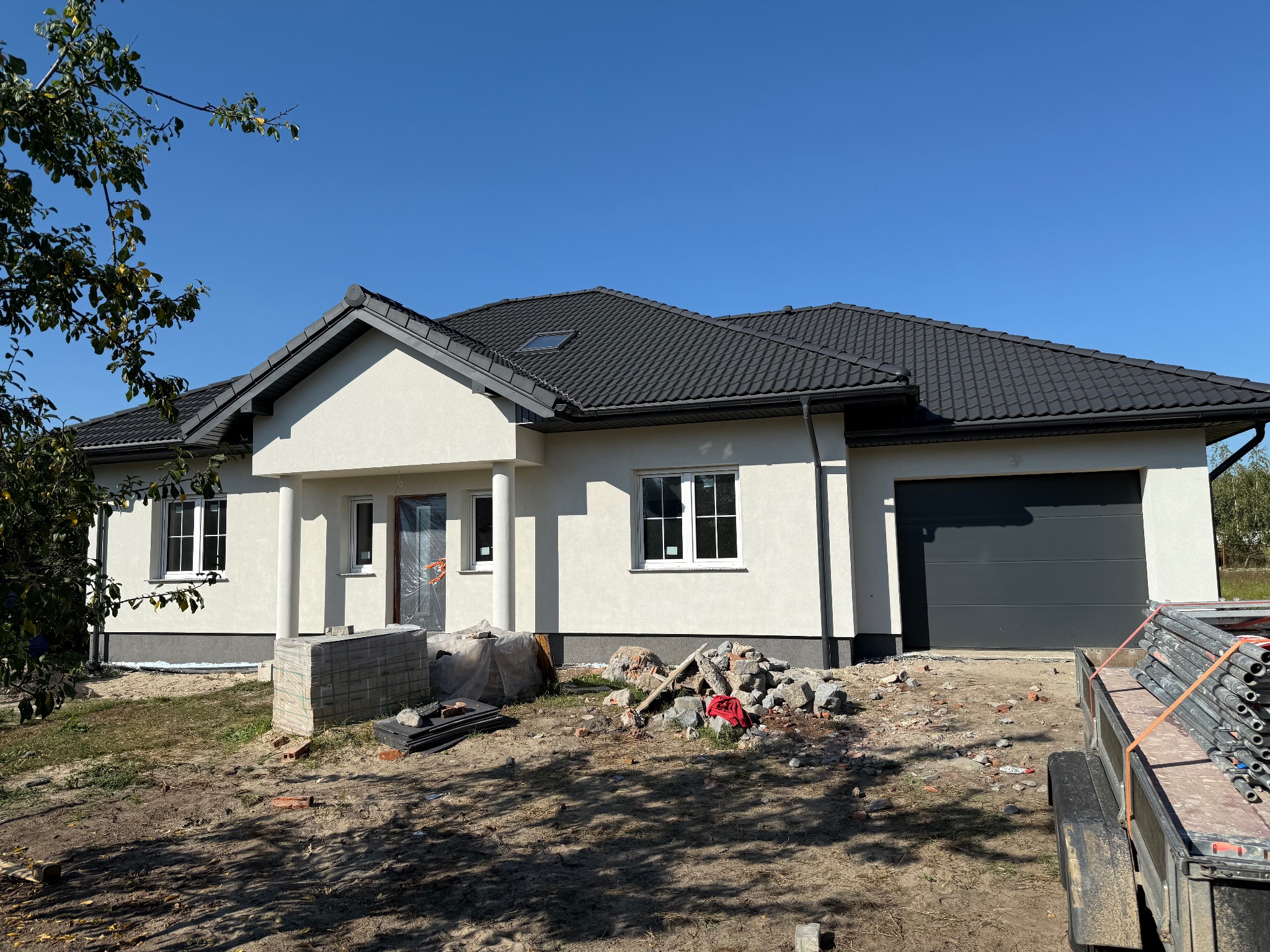 Nowoczesny dom jednorodzinny z grafitowym dachem i garażem, w trakcie prac wykończeniowych. Widoczne materiały budowlane i rusztowanie na przyczepie.