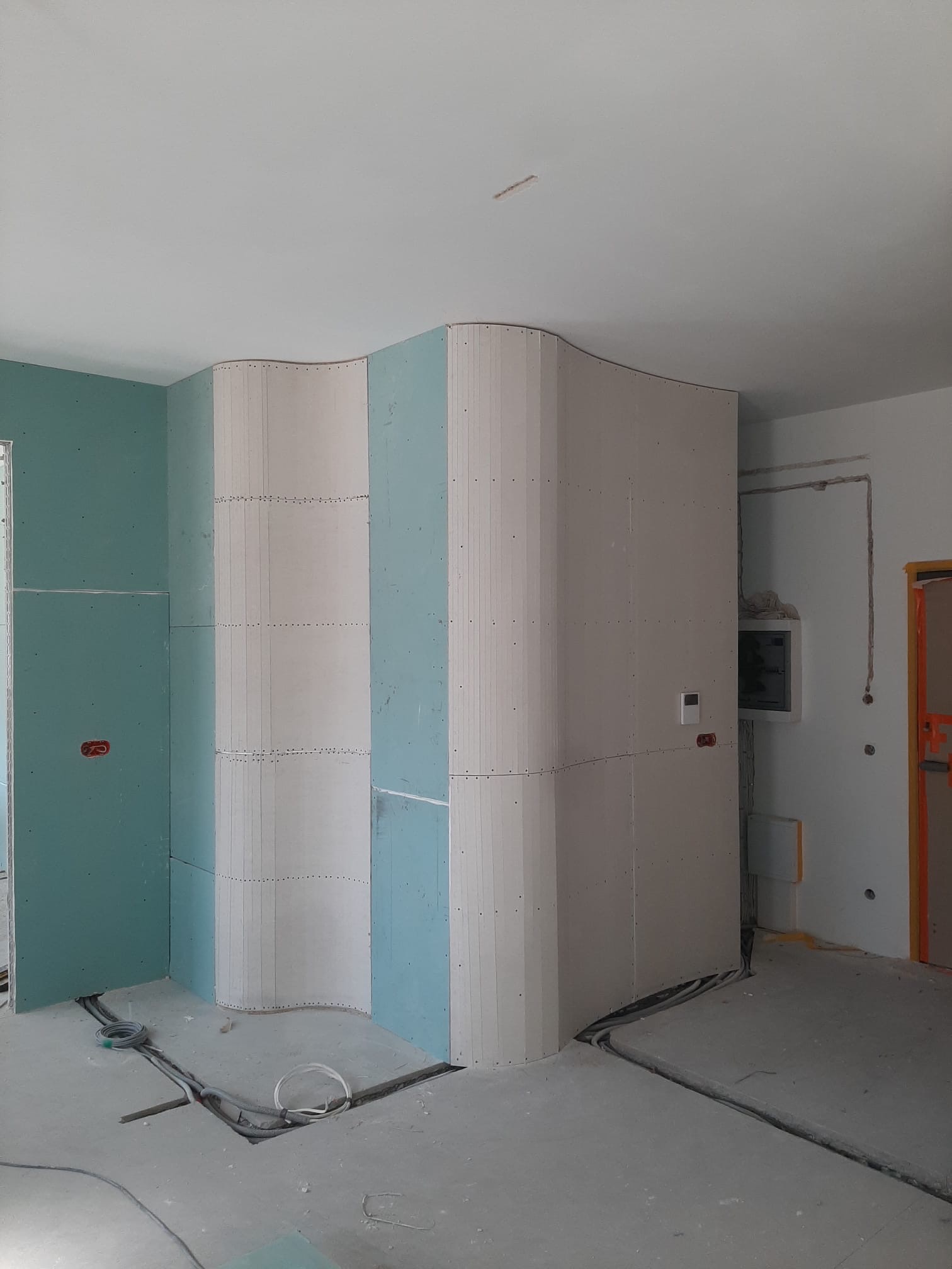 Narożnik z płyt kartonowo-gipsowych o falistym kształcie, w surowym wnętrzu. Widoczne przewody elektryczne w podłodze i skrzynka elektryczna na ścianie.