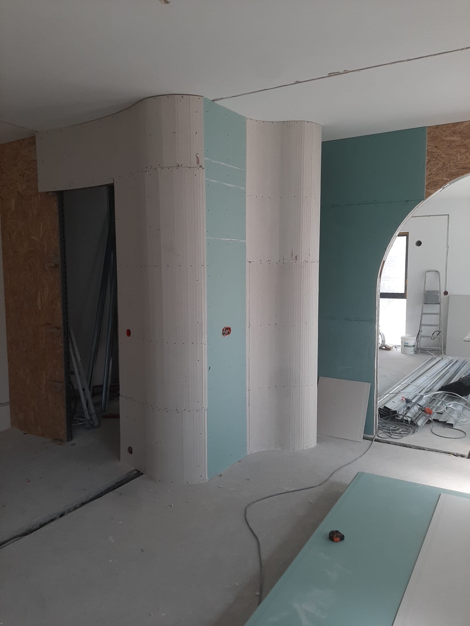 Nietypowa konstrukcja z płyt gipsowo-kartonowych w kształcie falistych kolumn, widoczne mocowania, surowy stan wykończenia wnętrza, elementy instalacji elektrycznej.