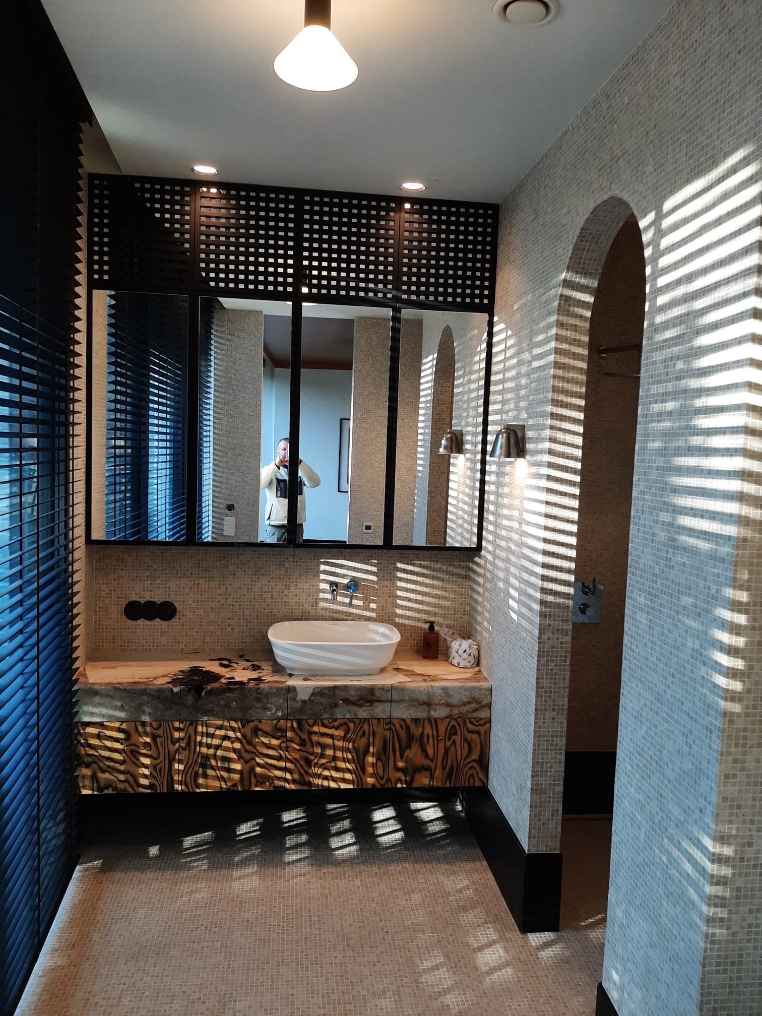 Nowoczesna łazienka z mozaiką, drewnianą szafką pod umywalką i lustrem z czarną kratką. Promienie słońca tworzą wzory na ścianach.