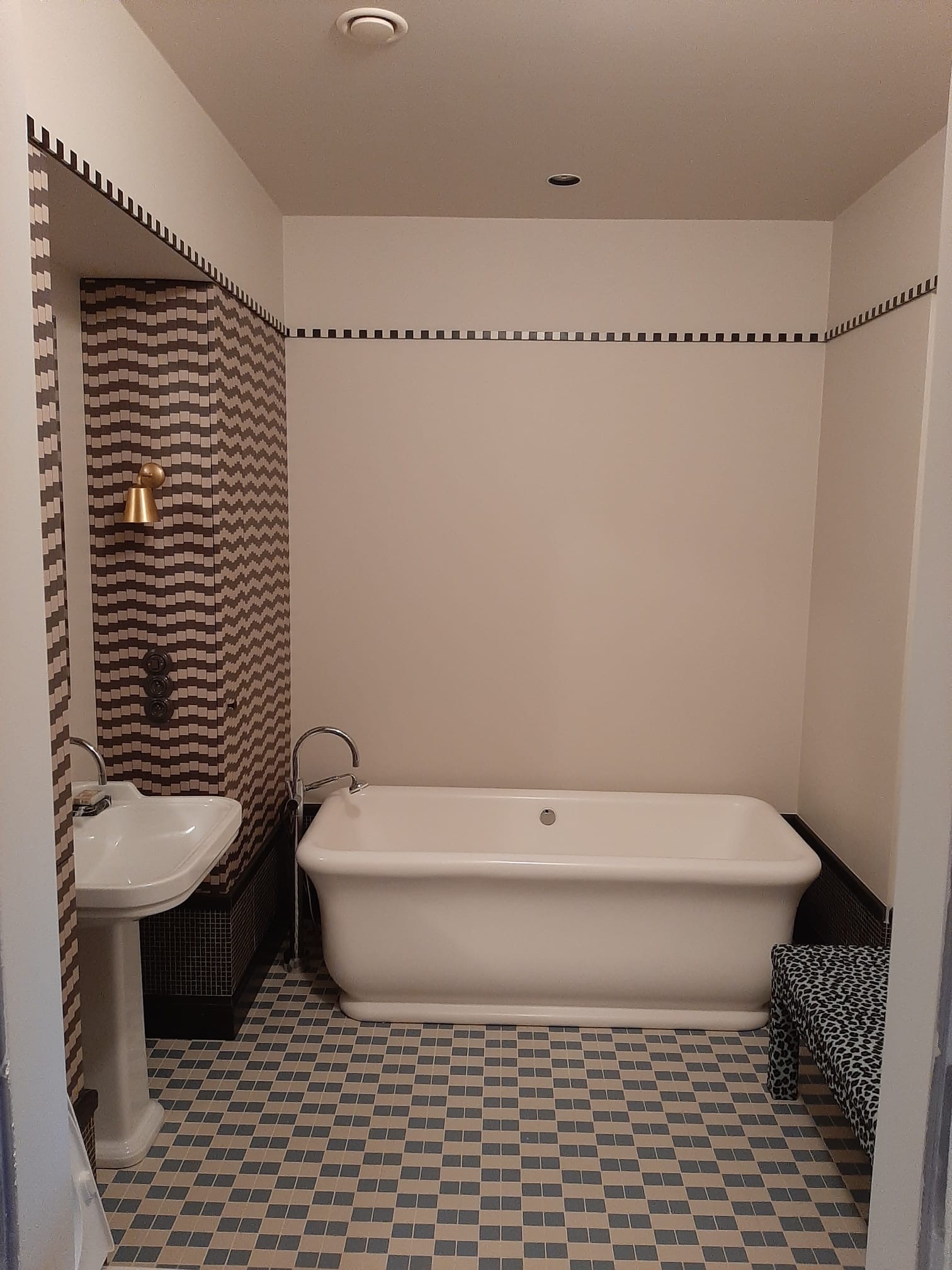 Elegancka łazienka w stylu retro z wolnostojącą wanną, umywalką na postumencie i wzorzystymi płytkami w odcieniach szarości i beżu. Dekoracyjne pasy na ścianach.