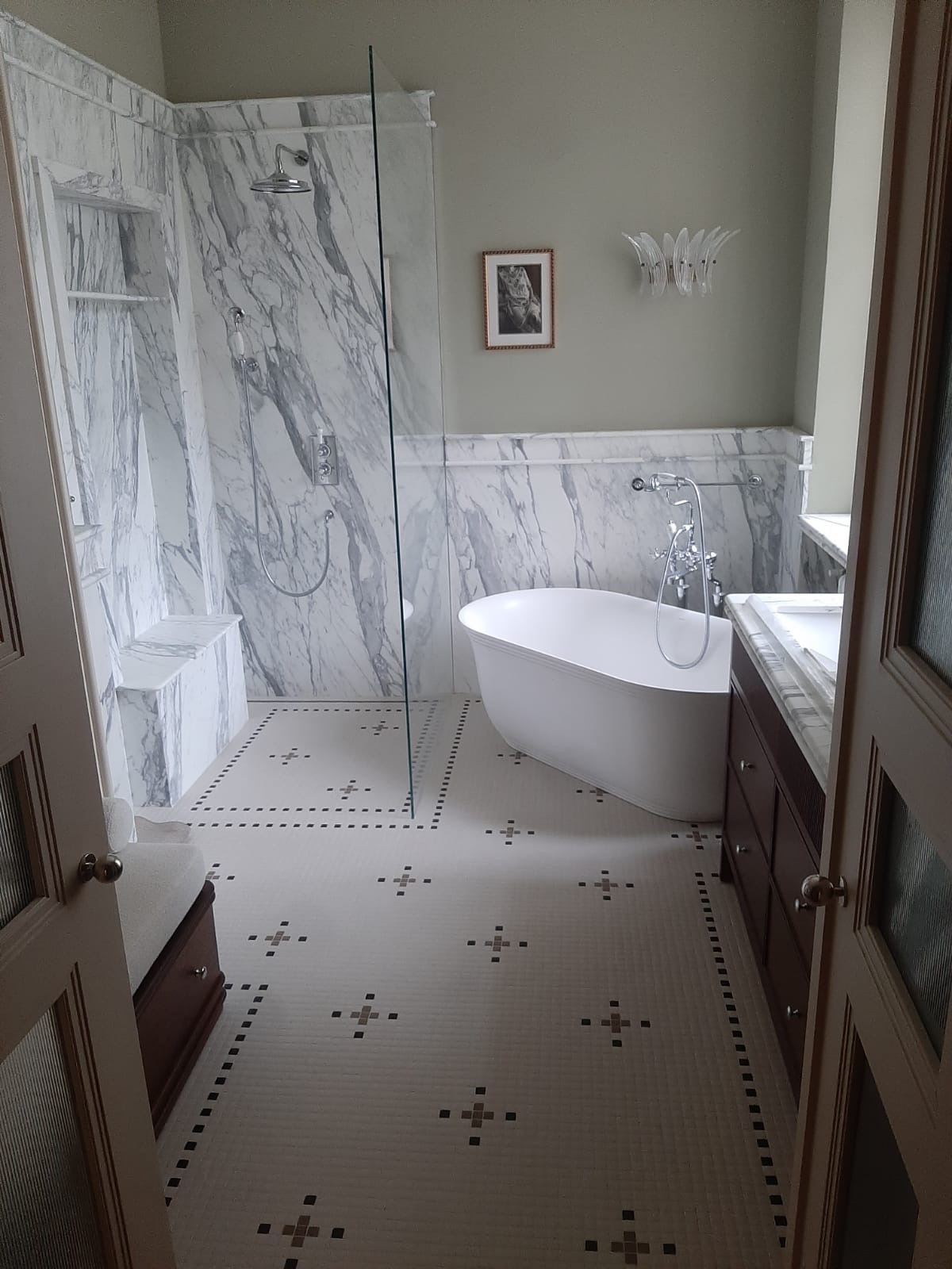 Elegancka łazienka z marmurowym wykończeniem ścian i kabiny prysznicowej, wolnostojąca wanna, mozaikowa podłoga i szafka z umywalką, widok zza uchylonych drzwi.