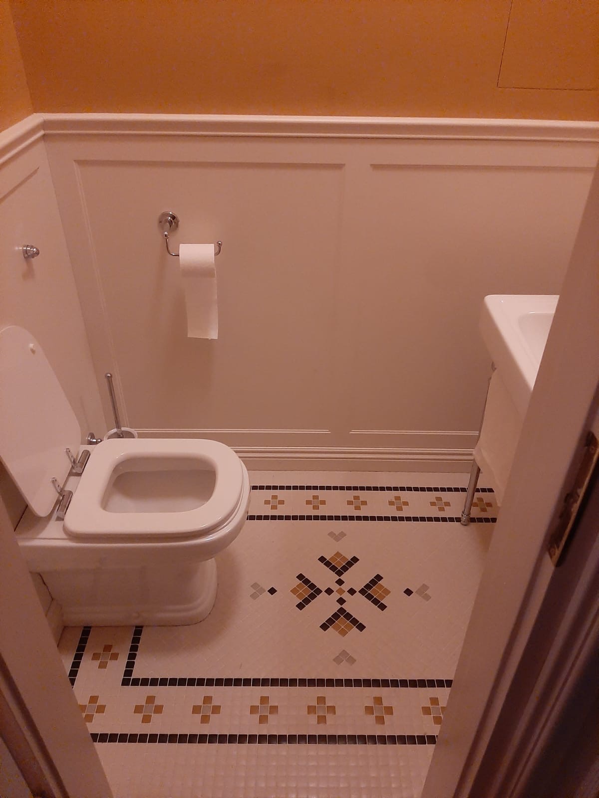 Wykończona łazienka z mozaikową podłogą w geometryczne wzory, białą toaletą i umywalką. Ściany wykończone panelami i pomalowane na jasny kolor.