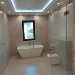 Nowoczesna łazienka z wolnostojącą wanną, kabiną prysznicową ze złotymi zawiasami i podświetlanym sufitem, wykończona płytkami w odcieniach beżu.