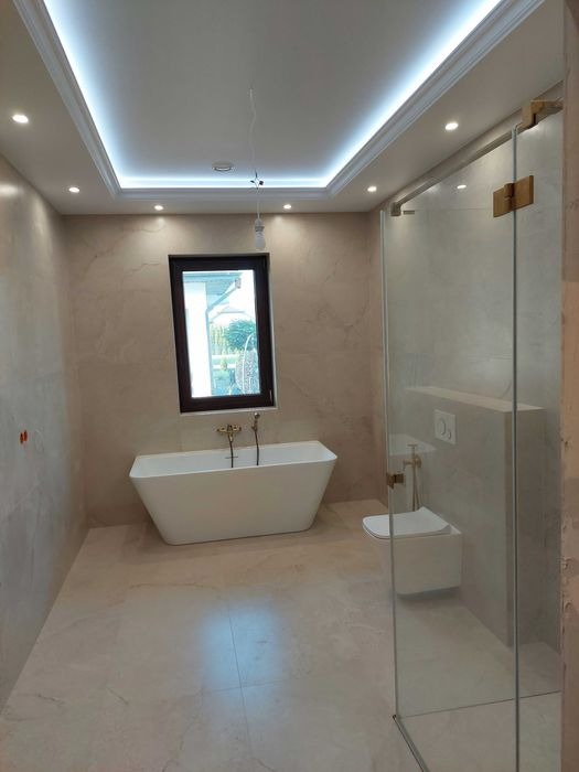 Nowoczesna łazienka z wolnostojącą wanną, kabiną prysznicową ze złotymi zawiasami i podświetlanym sufitem, wykończona płytkami w odcieniach beżu.