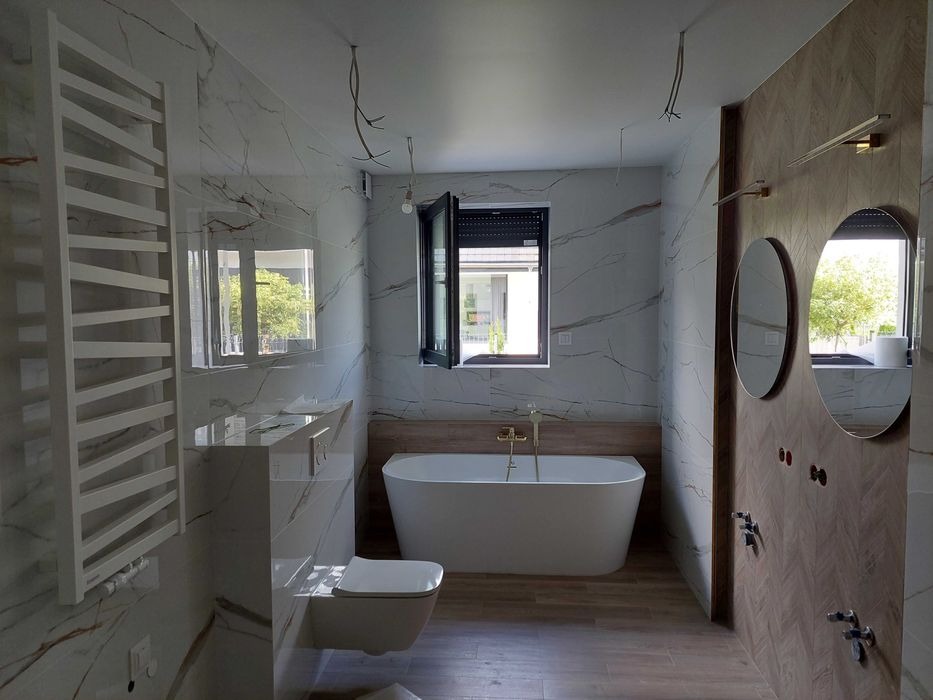 Nowoczesna łazienka z marmurowymi płytkami, wolnostojącą wanną z złotą armaturą i dwoma okrągłymi lustrami na ścianie z drewnopodobnym wzorem, widoczne niedokończone podłączenia oświetlenia...