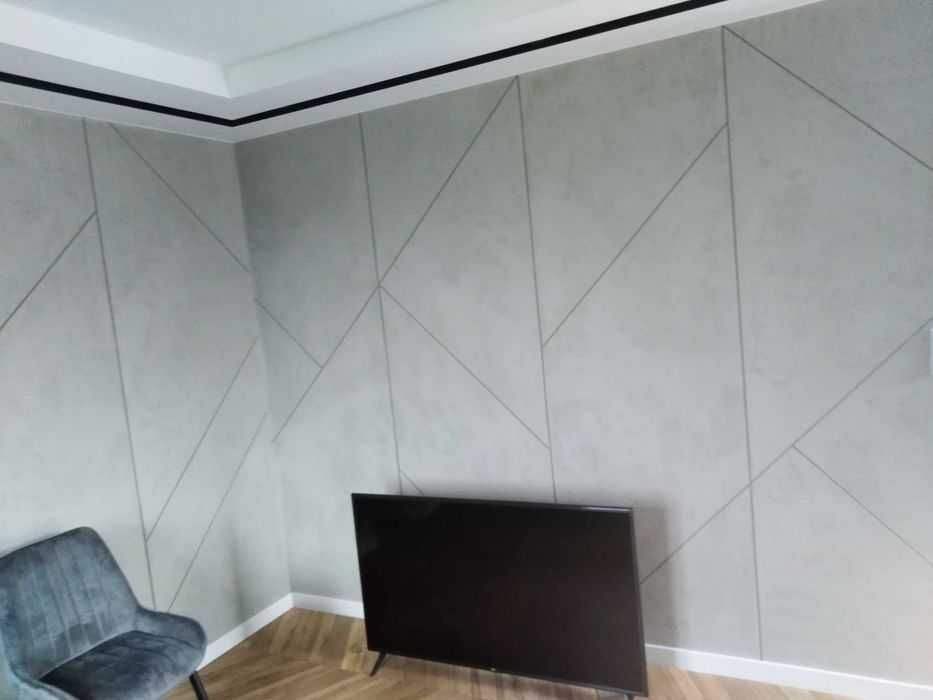 Szara ściana z geometrycznym wzorem z gładzi, czarny telewizor i fotel w rogu pokoju.