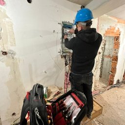 LUCA-ELEKTRO - Elektryk w niebieskim kasku pracuje nad rozdzielnicą elektryczną w surowym stanie budynku; otwarty plecak z narzędziami na pierwszym planie.