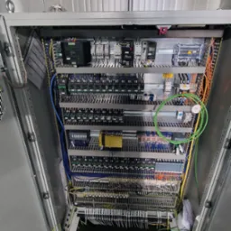 Otwarta szafa sterownicza z rozbudowanym okablowaniem, widoczne liczne moduły, przewody w różnych kolorach i listwy zaciskowe, na górze siatka kablowa