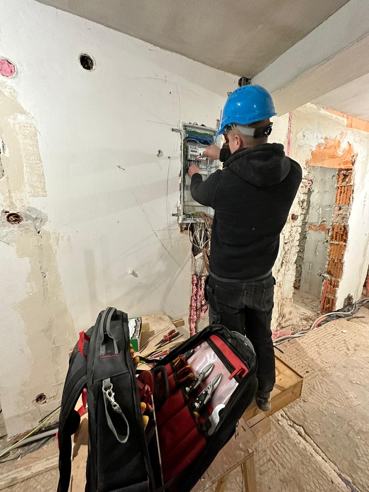 Elektryk w niebieskim kasku pracuje nad rozdzielnicą elektryczną w surowym stanie budynku; otwarty plecak z narzędziami na pierwszym planie.