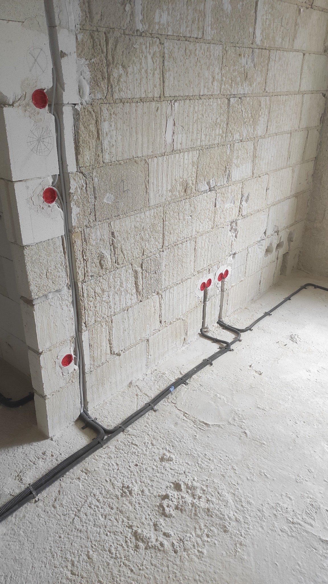 Szare rury karbowane prowadzone po podłodze i ścianie z surowej cegły, z czerwonymi puszkami instalacyjnymi w ścianie, na podłodze resztki zaprawy.
