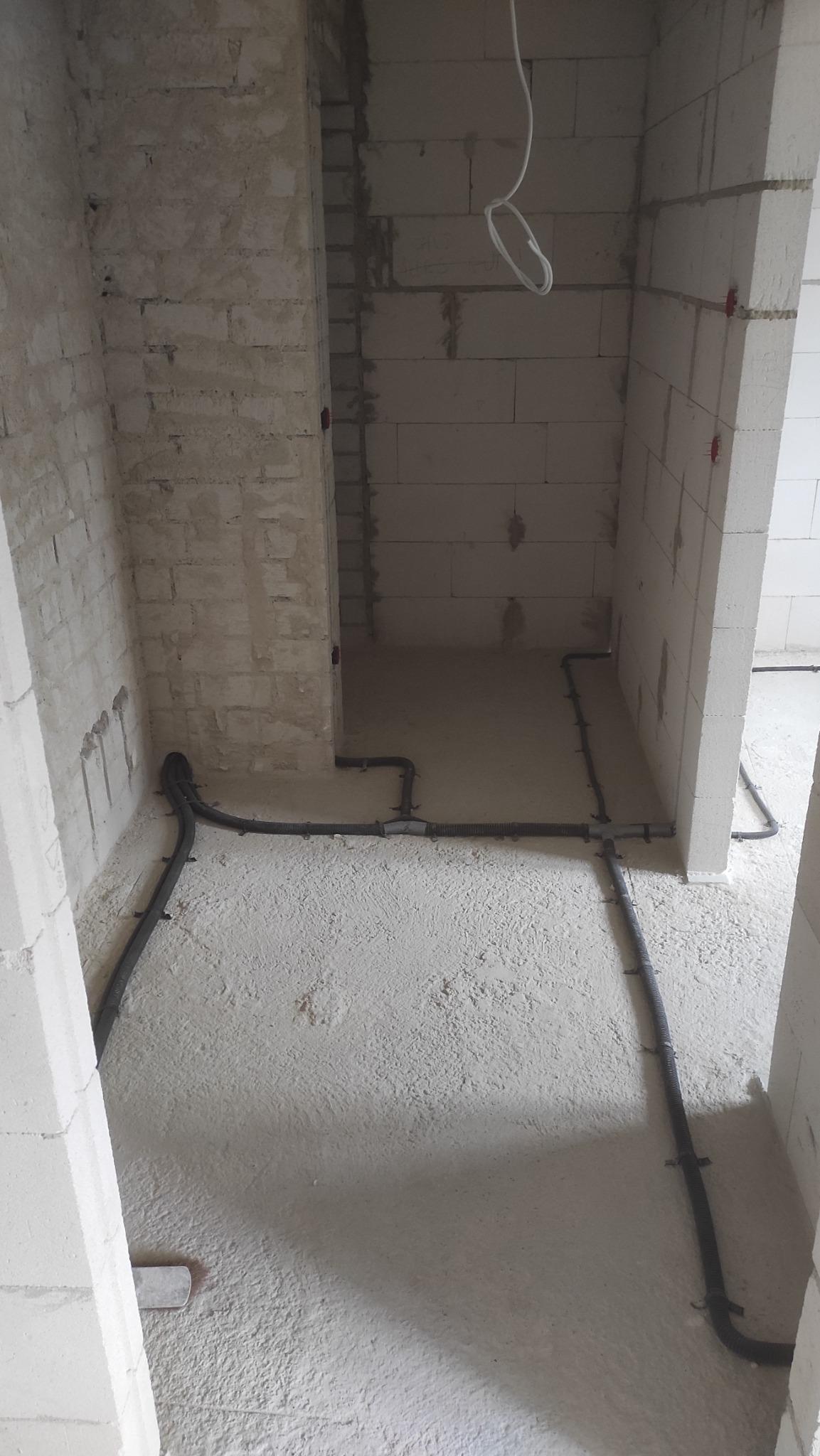 Szare rury instalacji elektrycznej prowadzone po podłodze i ścianach w surowym, niewykończonym pomieszczeniu z białych bloczków.