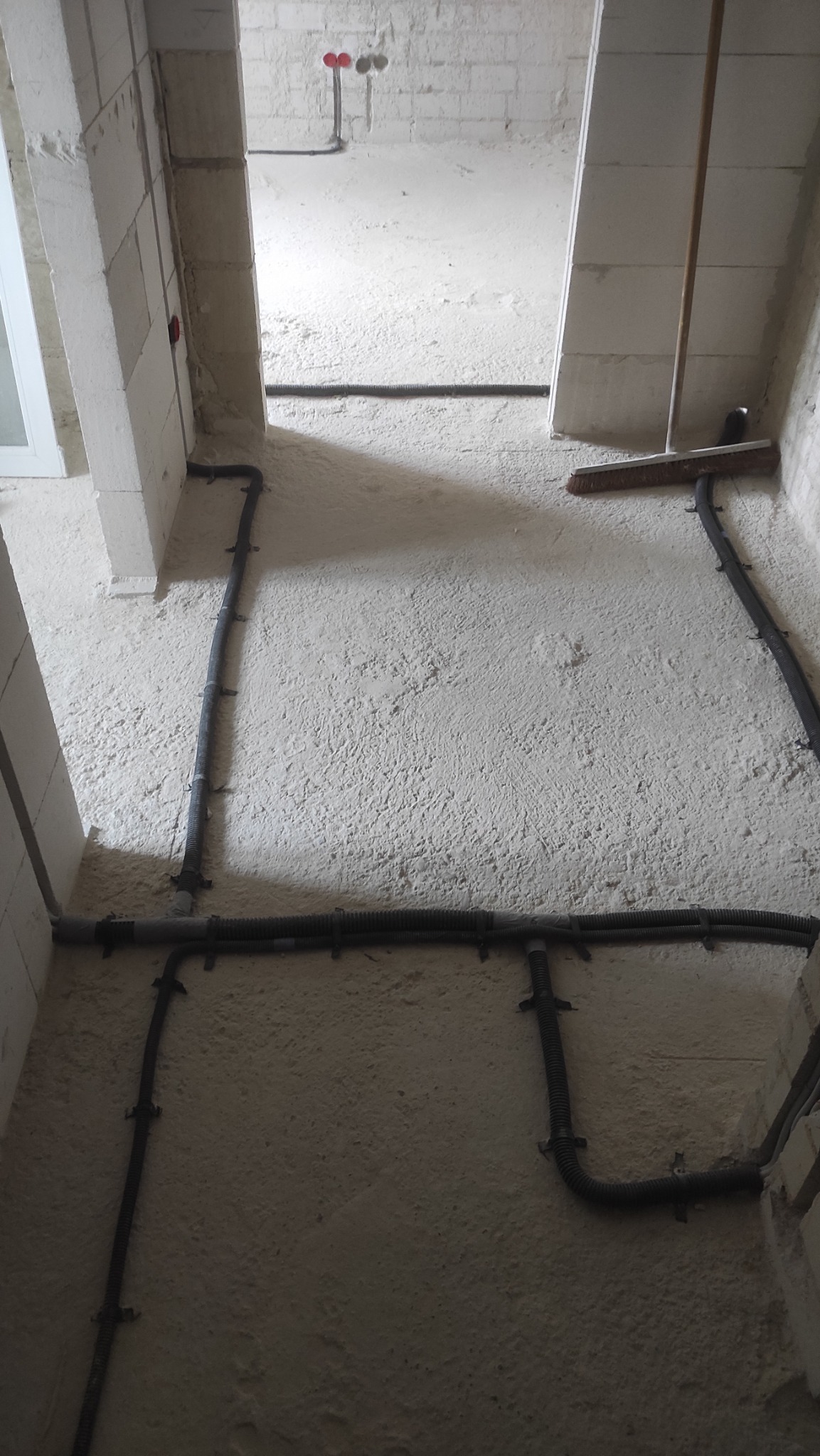 Wewnętrzne korytarze w surowym stanie z widocznymi przewodami elektrycznymi przymocowanymi do ściany, w tle wystające rury z instalacji wodnej i szczotka oparta o ścianę.