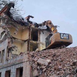 Jakub Kędzierski - Żółta koparka gąsienicowa CAT na szczycie sterty gruzu, rozbierająca częściowo zburzony, kilkupiętrowy budynek o jasnej elewacji z widocznymi zniszczeniami, na tle pochmurnego nieba i drzewa bez...