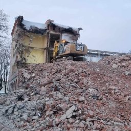 Jakub Kędzierski - Żółta koparka marki CAT na szczycie sterty gruzu, w tle częściowo zburzony budynek z odsłoniętymi żółtymi ścianami i fragmentami instalacji, drzewa bez liści i budynek w oddali.