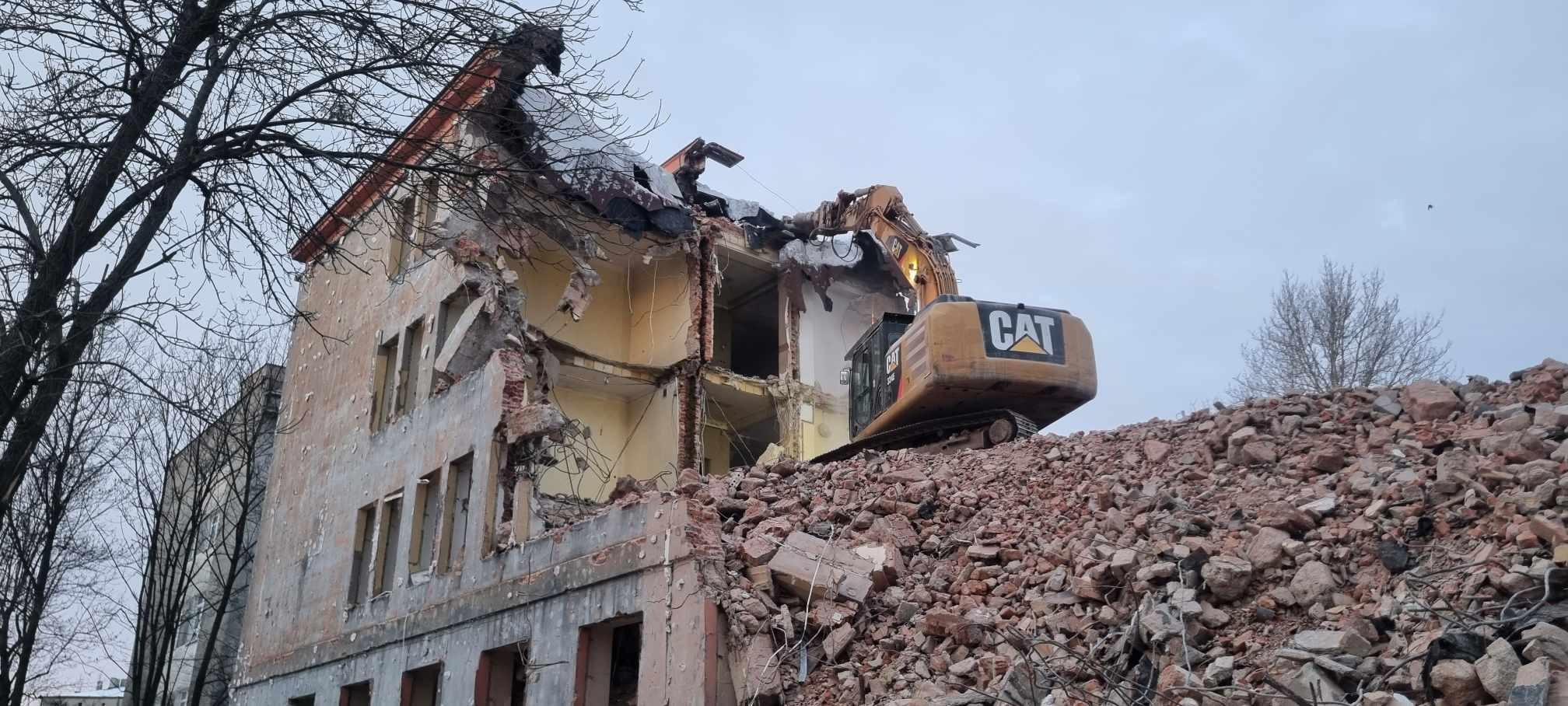 Żółta koparka gąsienicowa CAT na szczycie sterty gruzu, rozbierająca częściowo zburzony, kilkupiętrowy budynek o jasnej elewacji z widocznymi zniszczeniami, na tle pochmurnego nieba i drzewa bez...