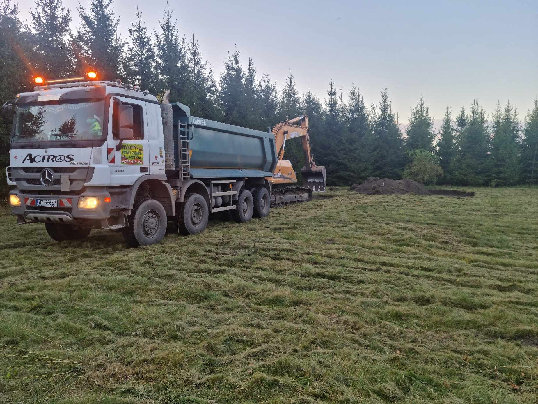 Ciężarówka wywrotka i koparka gąsienicowa pracują na zalesionym terenie, świeżo wykopany rów i pryzma ziemi, widoczny numer rejestracyjny ciężarówki.