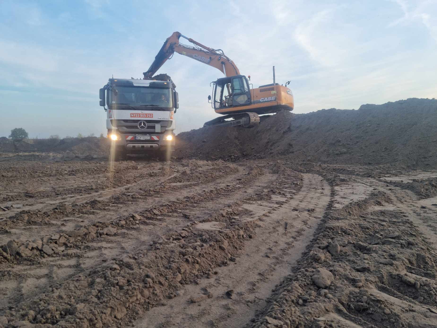Pomarańczowa koparka CASE na kopcu ziemi obok białej ciężarówki Mercedes z włączonymi światłami, na placu budowy z widocznymi śladami opon na ziemi.